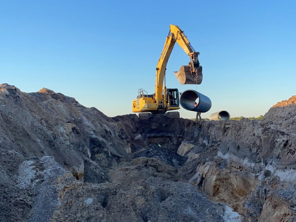 Excavator lifting a large concrete storm-water pipe into a deep dirt pit — Marathon, Florida